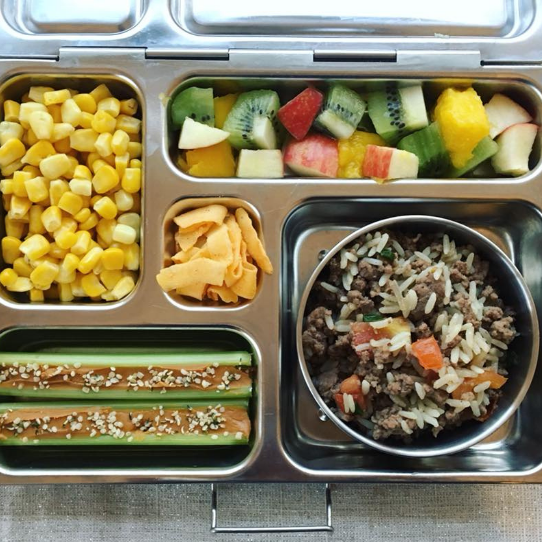 Main image for the article [The Ultimate Lunch Packing Guide]. Pictured is an open lunchbox with mixed fruit, corn, rice, hamburger and tomatoes, and celery with peanut butter on top.