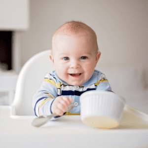 Main image for the article [Feeding Schedules (6-12 months)]. Pictured is a baby sitting in a highchair, playing with a spoon and bowl. 