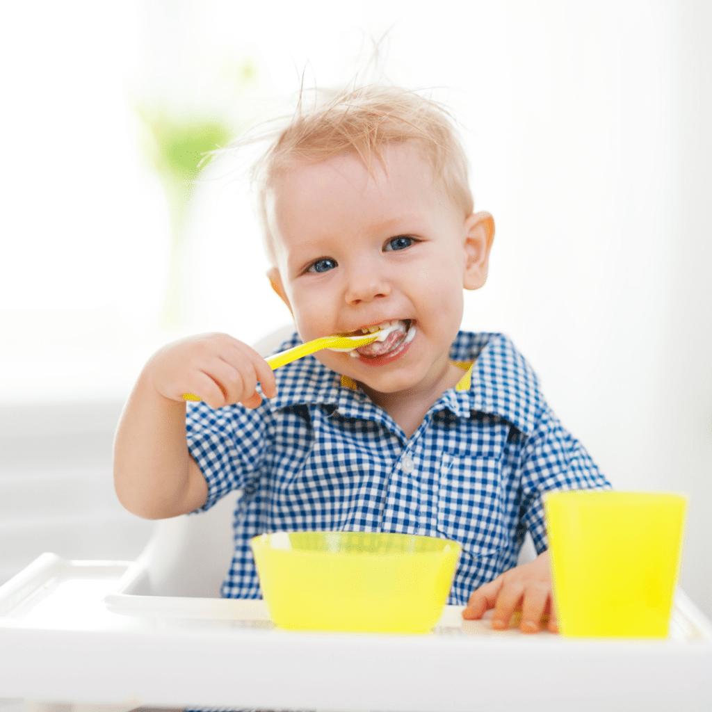 Main image for the article [How to teach mindful eating to your toddler]. Pictured is a toddler sitting at a table eating yogurt with a spoon.