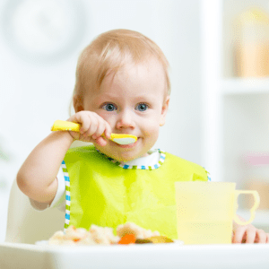 Main image for the article [How to Help Your Toddler Sit (and Stay) at the table].  Pictured is a toddler eating a plate of food in their highchair. 