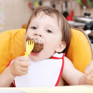 Main image for the article [How to Serve Meat to Babies/Toddlers]. Pictured is a toddler eating a piece of meat with a fork.