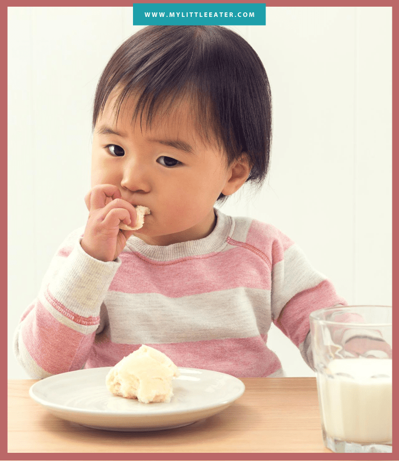 toddler in a pink and white striped sweater eating a dinner roll; half is still on the plate, and a glass of milk beside it on the table