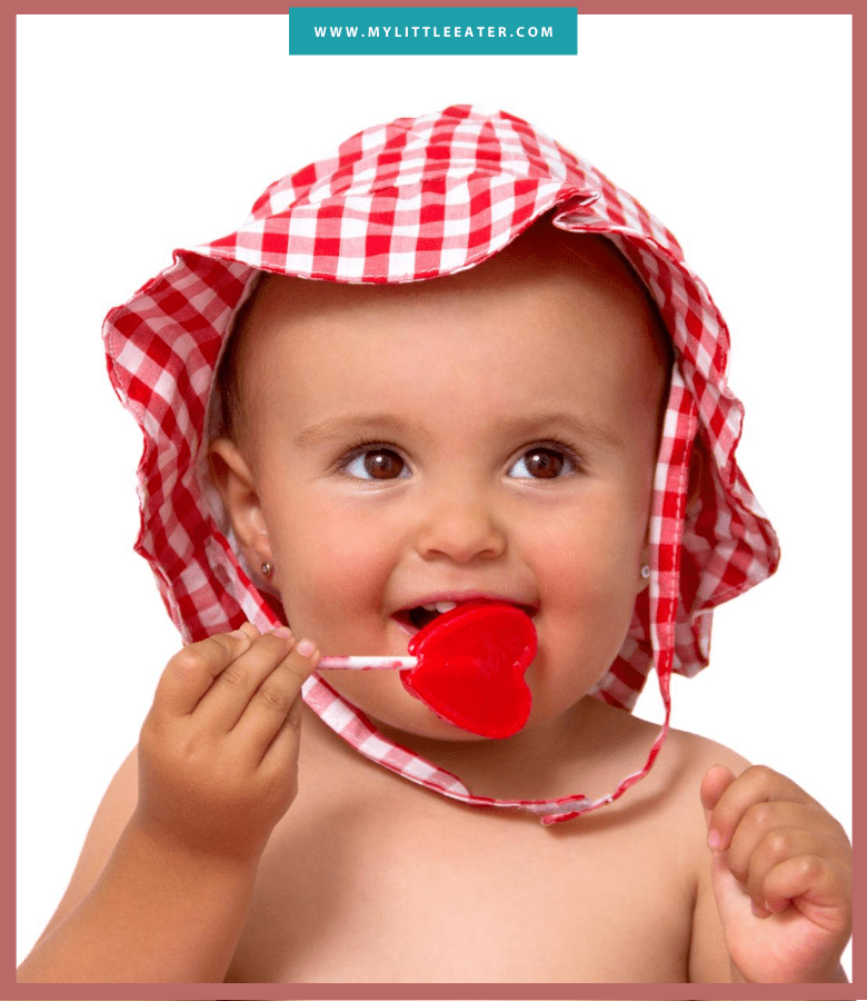 white baby in red sunhat eating a heart shaped lollipop