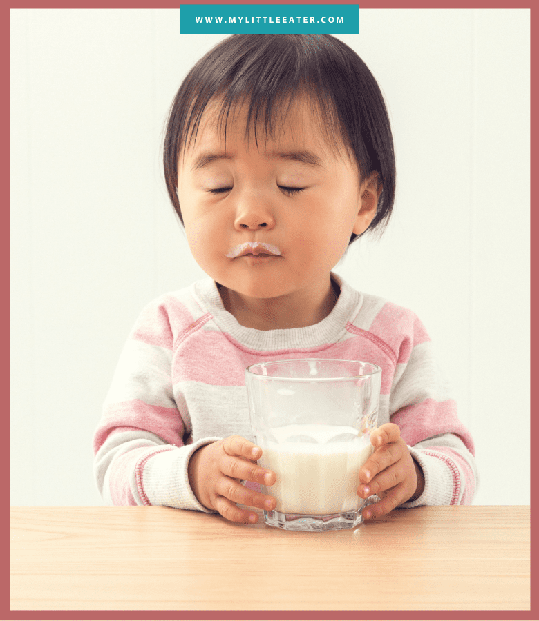 A toddler sits at a table with her eyes closed, holding a glass of milk. The toddler has a "milk moustache" from drinking the milk. Image has a maroon border, and the website "mylittleeater.com" is printed at the top in white font on a aqua background.