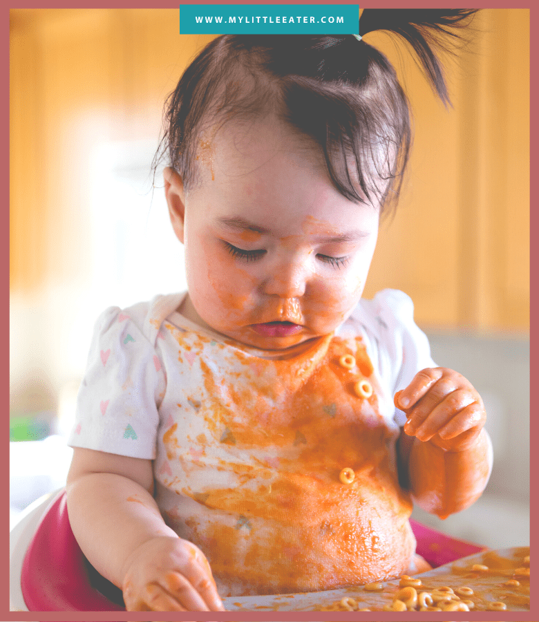 A baby sitting in a high chair eating with food all over her table, face, and onesie.