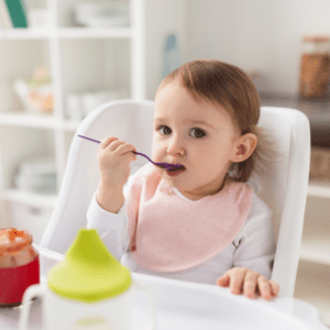 Main image for the article “How to Transition to Finger Foods When Your Baby is Stuck on Purees”. Pictured is a little girl sitting in her highchair, eating jarred puree baby food with a spoon.