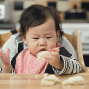 Episode art for episode "#43: The Best Bread for Baby Led Feeding." Pictured is a baby sitting in a high chair looking at a piece of bread.