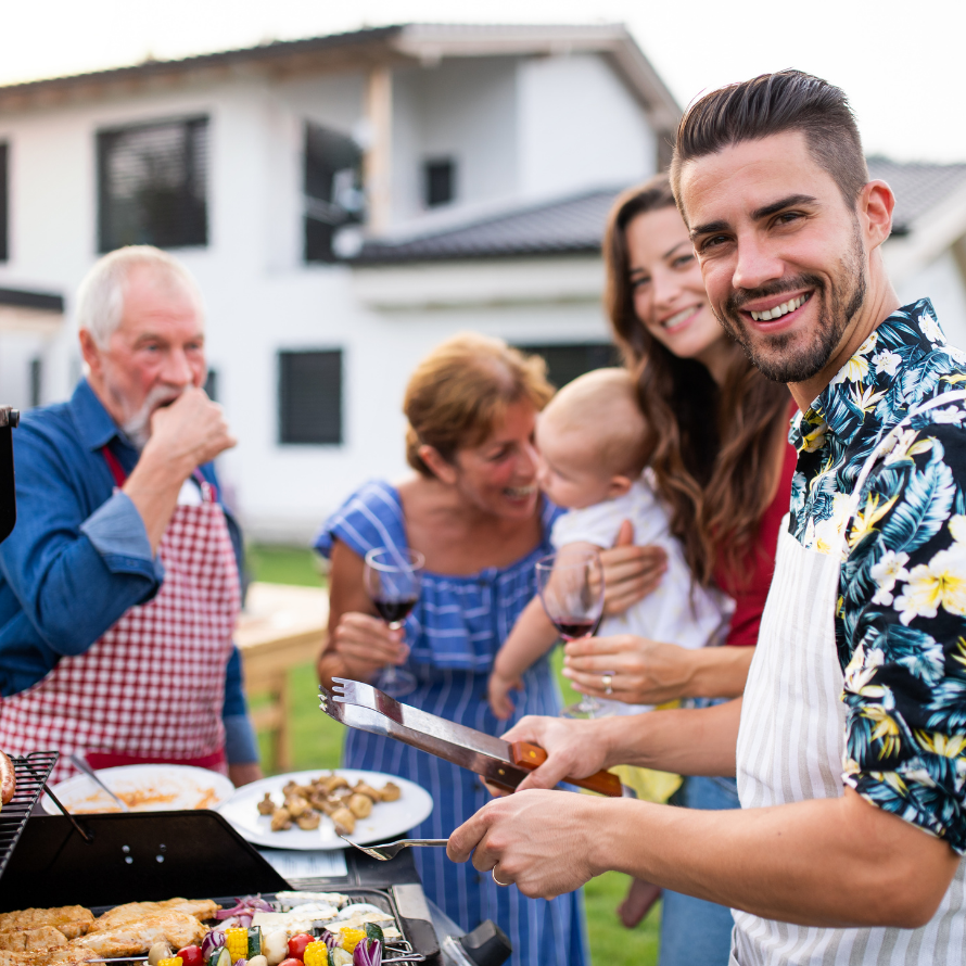 Featured image for the article: "BBQ Food and your baby - What you NEED to know". Pictured is a family gathered around the barbecue, including parents, grandparents, and young baby.