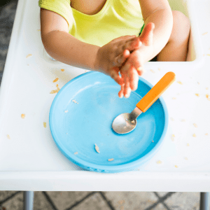 Featured image for the article: "The best plates for toddlers and picky eaters: divided or undivided?". Pictured is an empty plate with a spoon on top of a high chair.