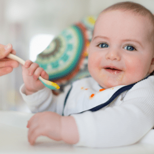 Baby happily eating puree baby food and trying to take the spoon from their mom.
