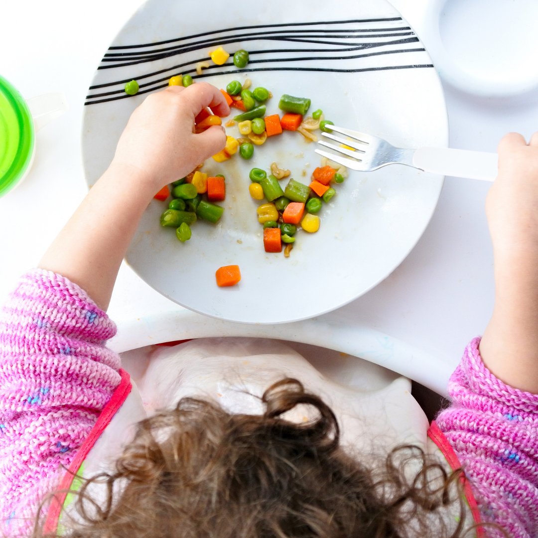 Toddler sitting in highchair eating carrots, corn and peas