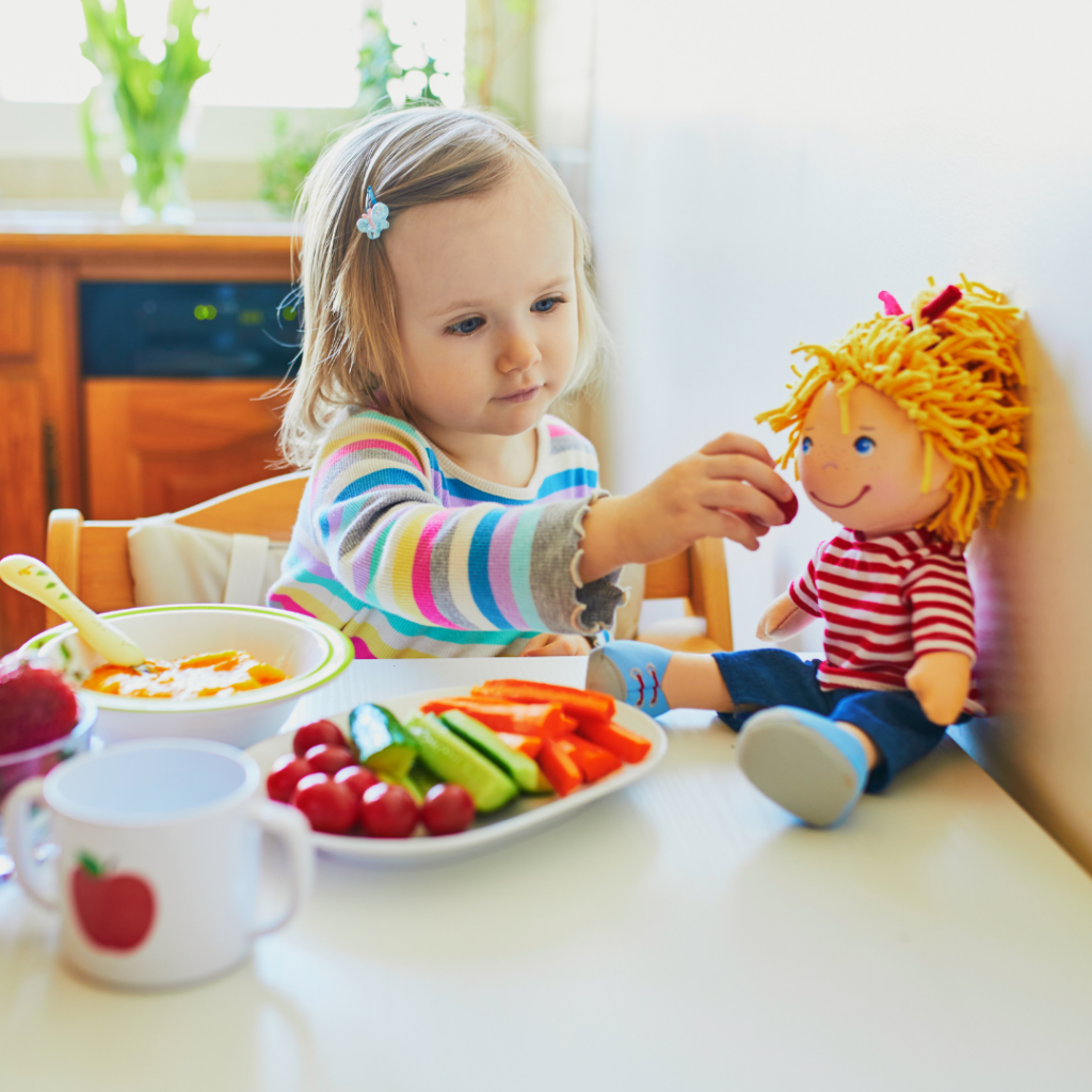 Toddler eating after daycare and feeding their doll with food and a cup on the table.