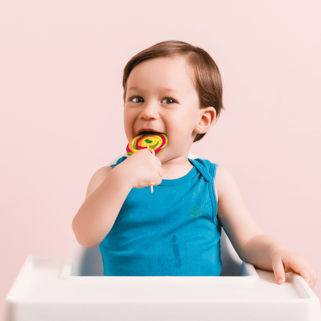 Happy toddler with a sweet tooth in a high chair eating a lollipop. 