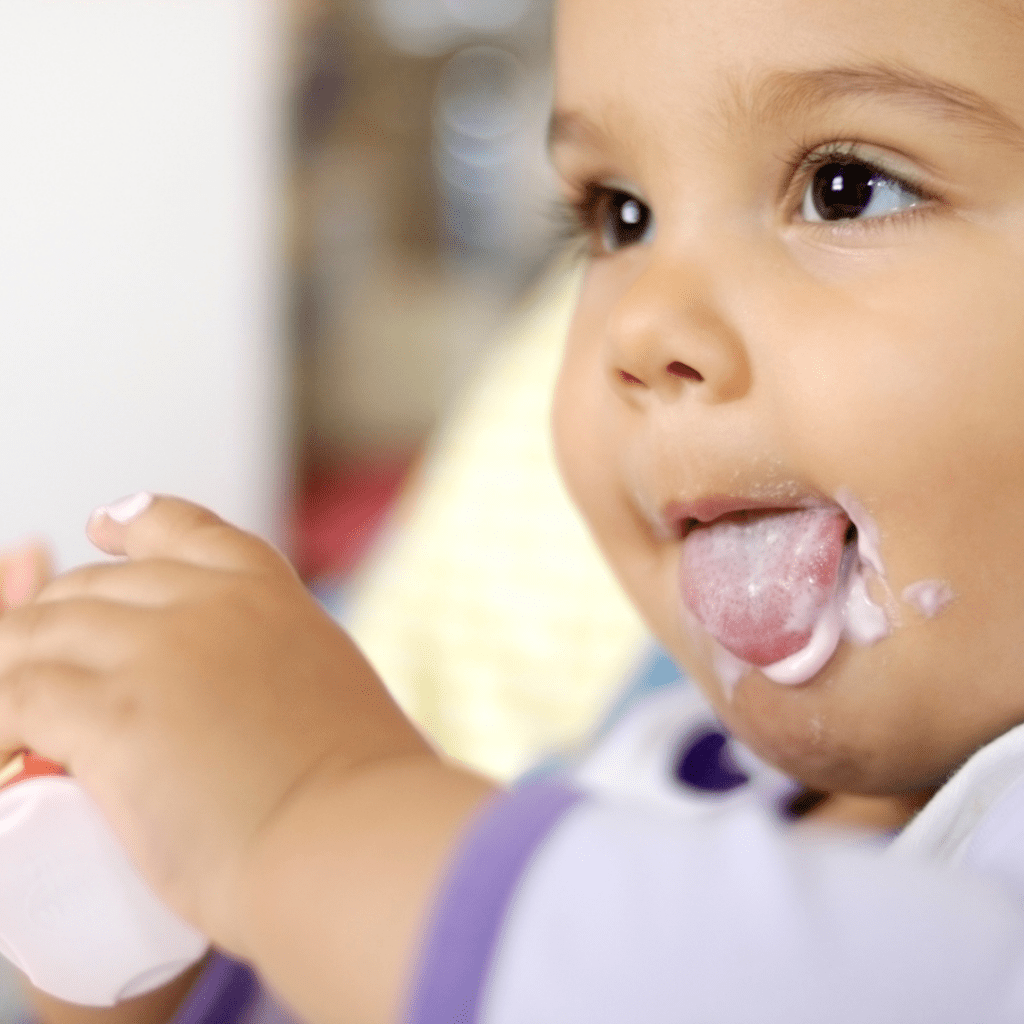 Babies can eat yogurt, introducing yogurt has many benefits for them; baby eating yogurt with some on their face.