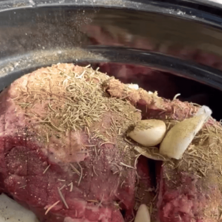 Lamb roast cooking in a crockpot with herbs, garlic, and onions.
