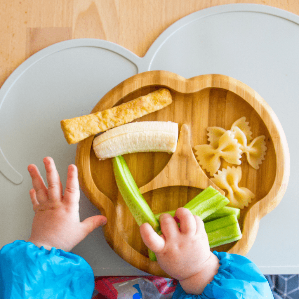 Various solid foods for baby led weaning on a plate with a baby reaching for them, including banana, pasta, omelet, and cucumber spears.
