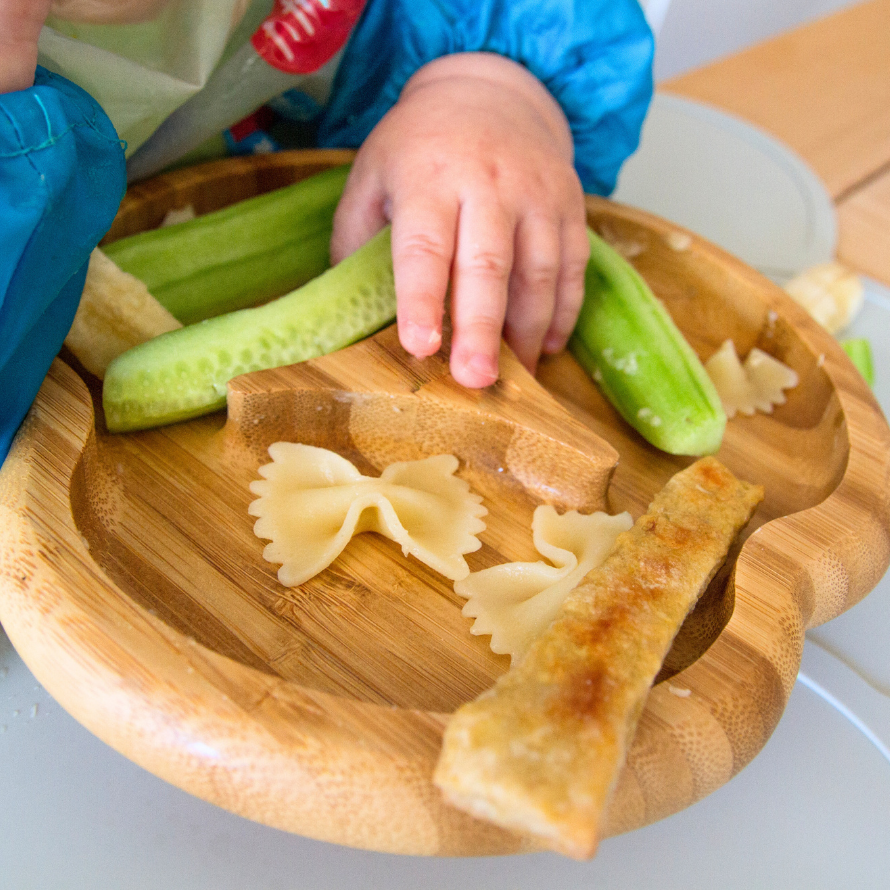 Introducing foods to baby on a bamboo plate, including pasta, cucumber, and toast.