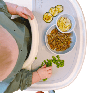 Baby in a high chair eating, baby led weaning style, zucchini, ground beef, mashed potatoes, and peas.