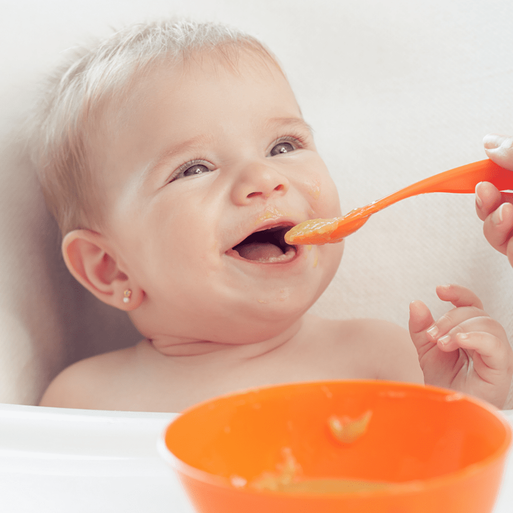 A baby smiling in a high chair, happily being fed a spoon of purees.