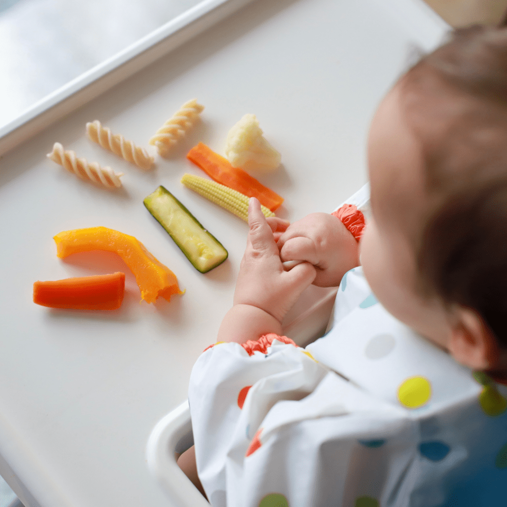 When can babies eat pasta; baby in a high chair with various foods in front of them, including peppers, cucumber, cauliflower, and rotini pasta.