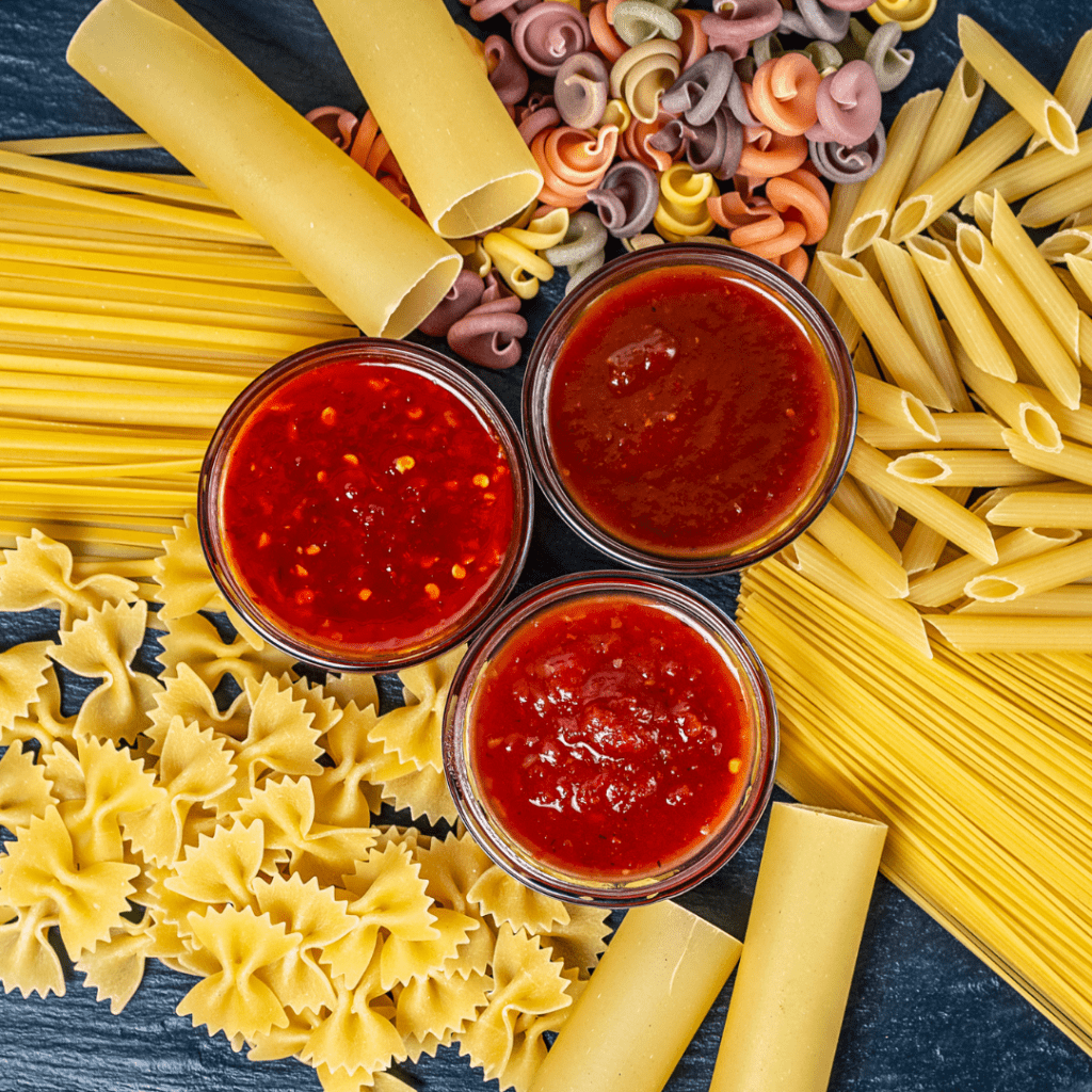 Various shapes of raw pasta spread around 3 small bowls, each containing a different sauce.