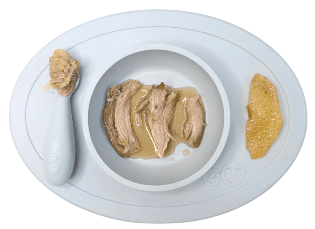 Slow-cooked lamb on a baby plate shown in strips for baby led weaning, mixed with mashed apple on a preloaded spoon, and with cooked apple on the side.