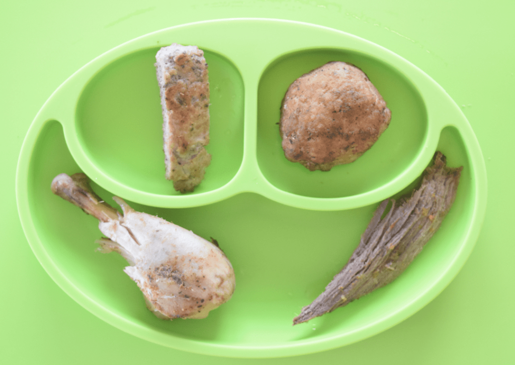 Green divided baby plate with fully cooked meat, including a chicken drumstick, meatball, and strips of meat.