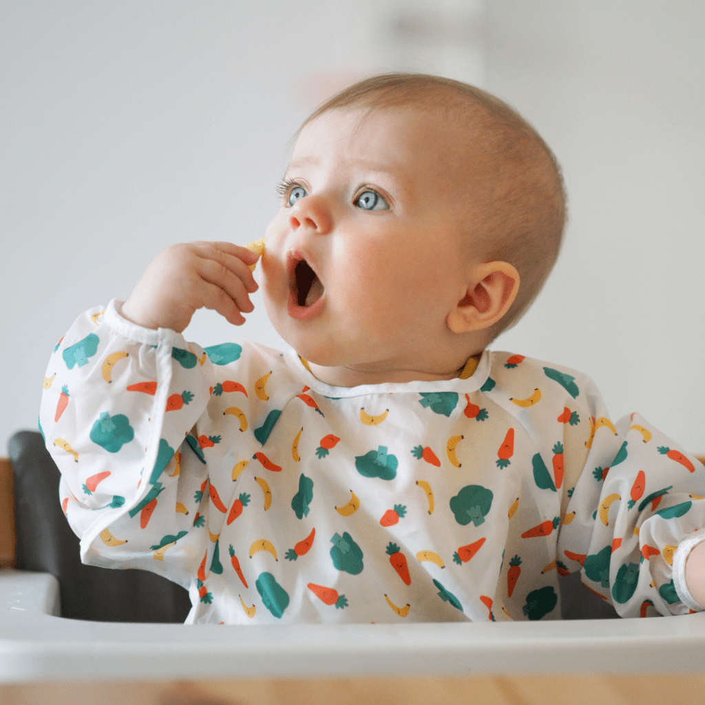 Baby sitting in high chair with mouth open wide, bringing a piece of food to their mouth using their developing pincer grasp.