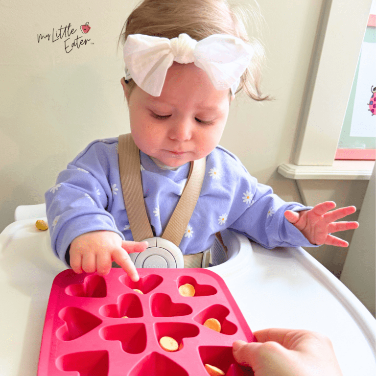 Baby sitting in high chair eating Amara Organic Smoothie Melts using her pincer grasp to take them out of a silicone baking tray with small heart compartments.