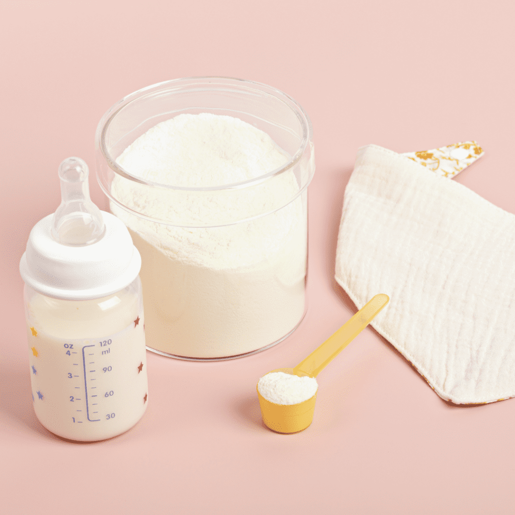 White bib, can of baby formula, baby formula in a yellow scoop, and baby bottle filled with milk on a pink background.