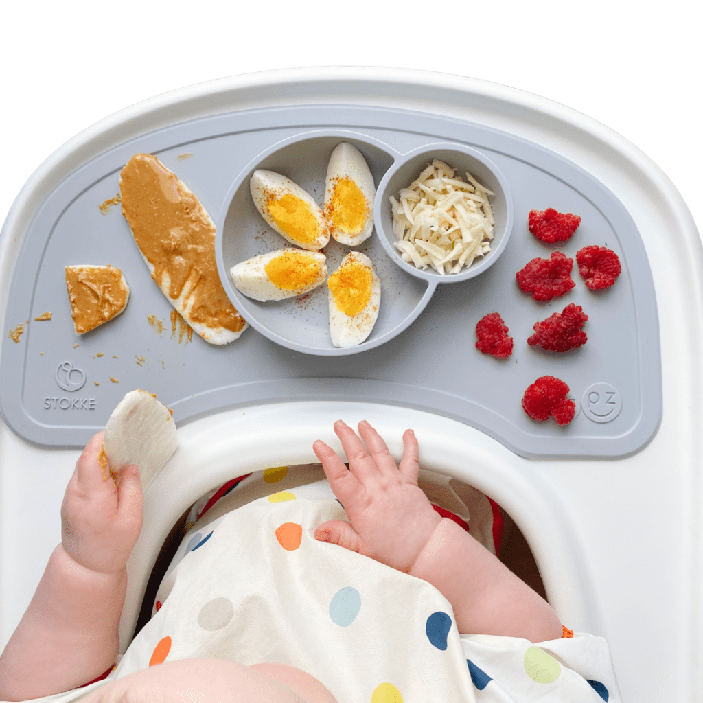 Overhead view of baby with finger foods on their highchair tray including boiled eggs with paprika, shredded cheese, rice rusks with nut butter, and squished raspberries.