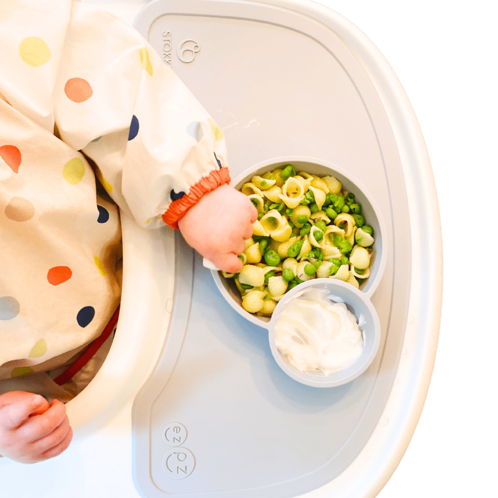 An overhead shot of a baby eating a bowl of shell-shaped pasta mixed with green peas and plain yogurt on the side.