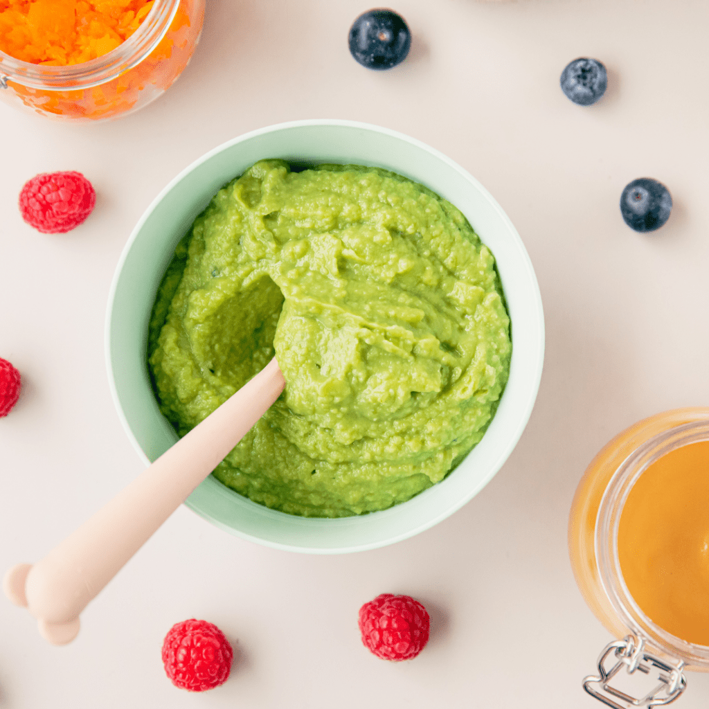 A baby's food (green puree) in a bowl with a spoon, surrounded by raspberries, blueberries, and jars of orange baby food.