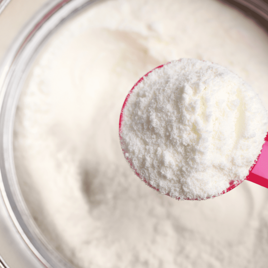 A pink scooping spoon full of powdered formula is held above a full container of baby formula.