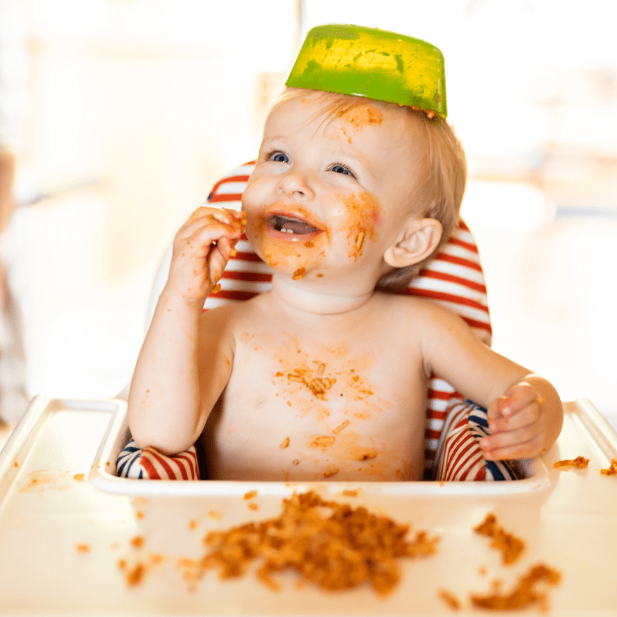 Baby making a mess while eating pasta in a high chair, has placed their bowl on their head.