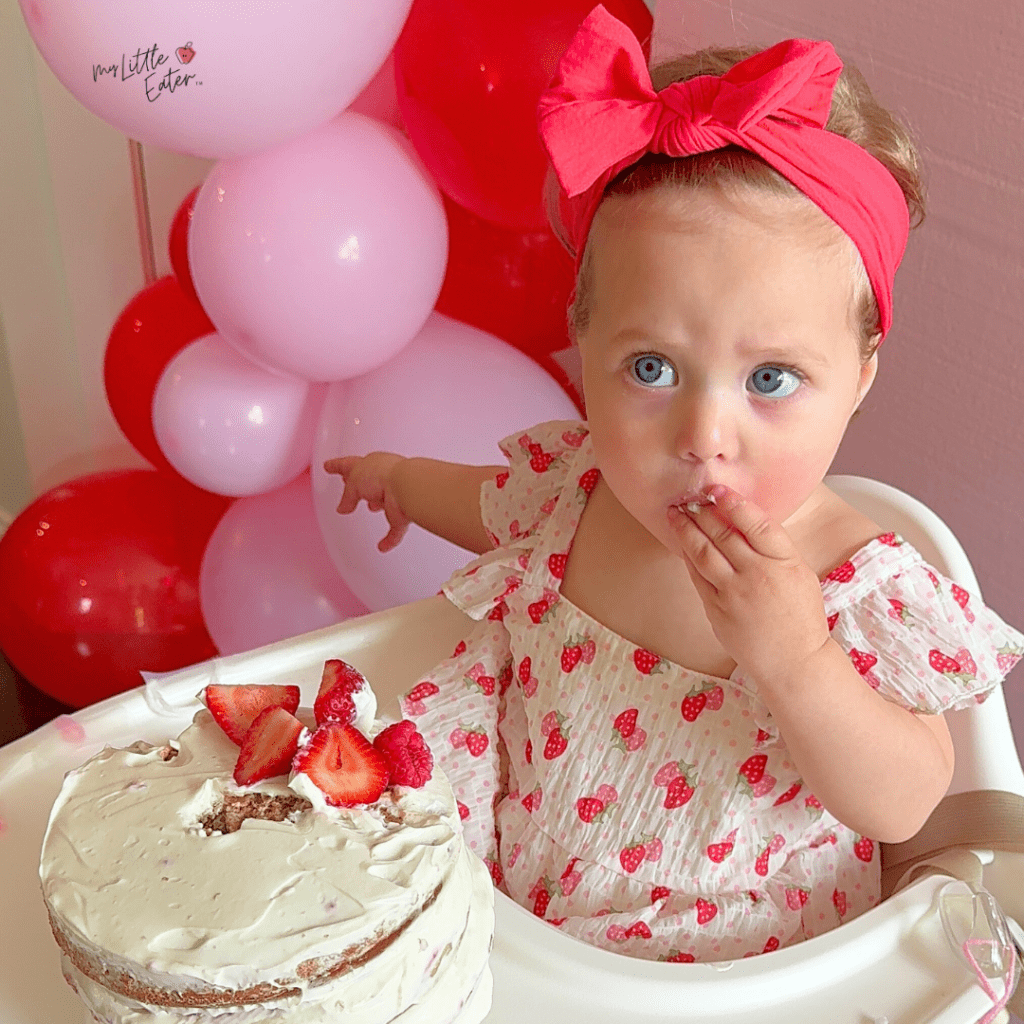 Baby sits with their strawberry cake decorated for their berry 1st birthday.