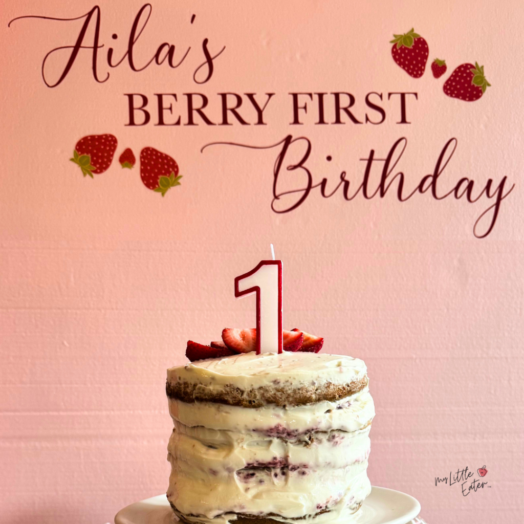 Aila's berry first birthday is written above a smash cake with a coconut, yogurt, or cream cheese frosting.