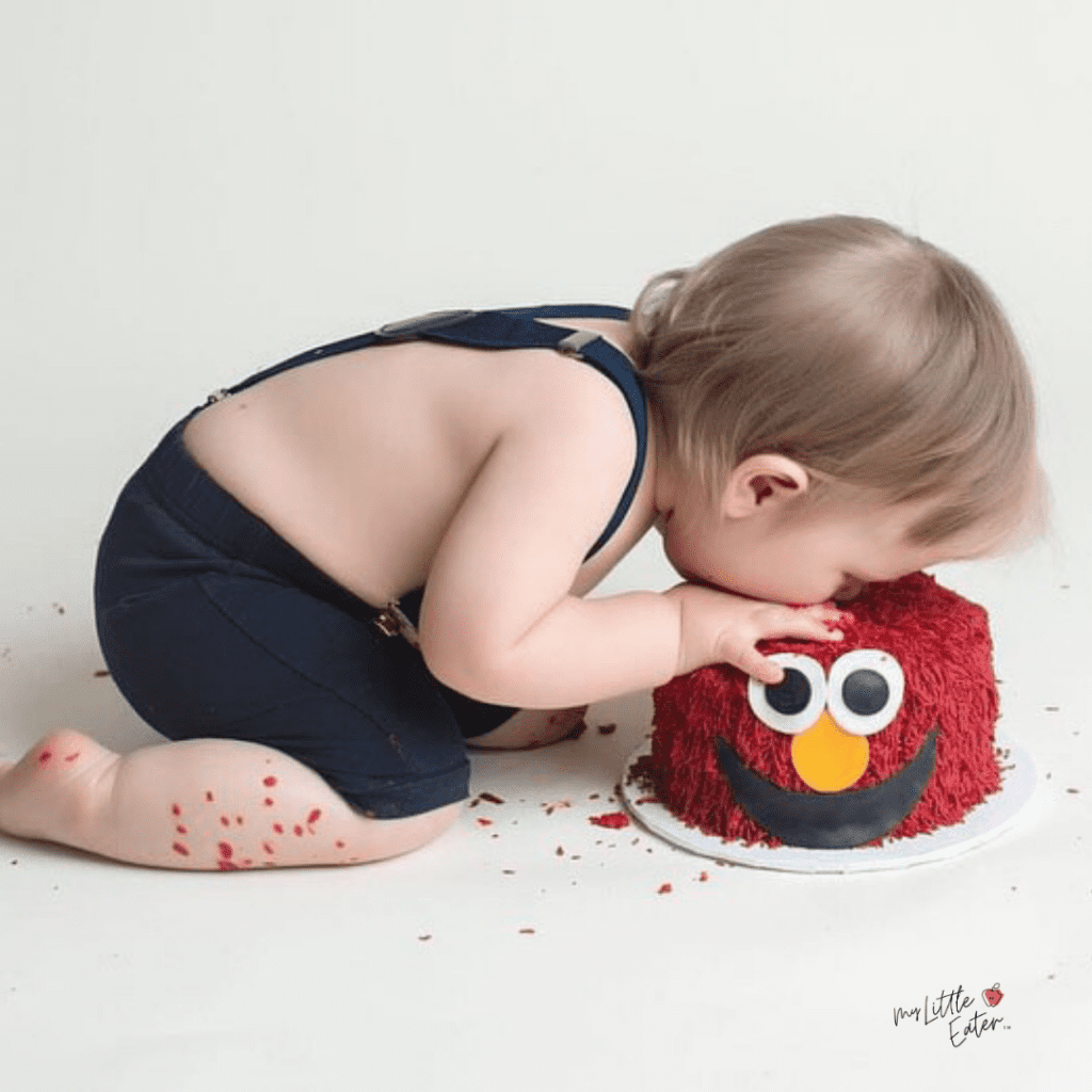 A baby puts their face into their smash cake that is decorated as Elmo.