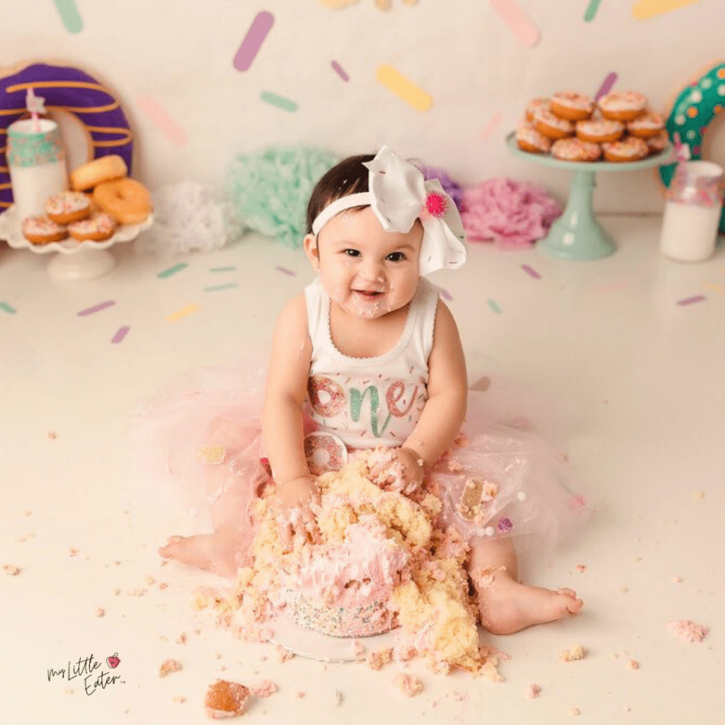 Baby smiles while sitting in a completely smashed cake for first birthday.
