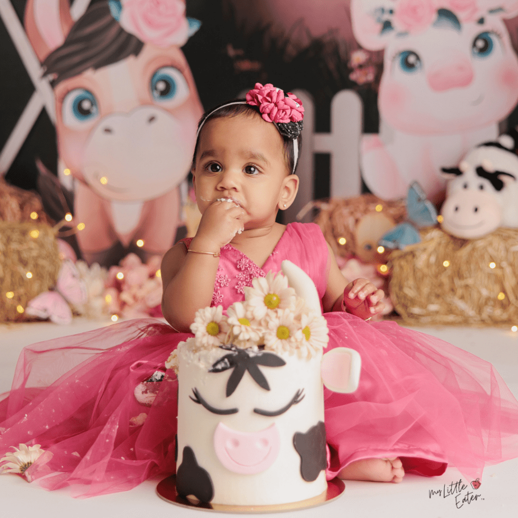 A baby in a pink dress sits behind their smash cake that is decorated as a cow.