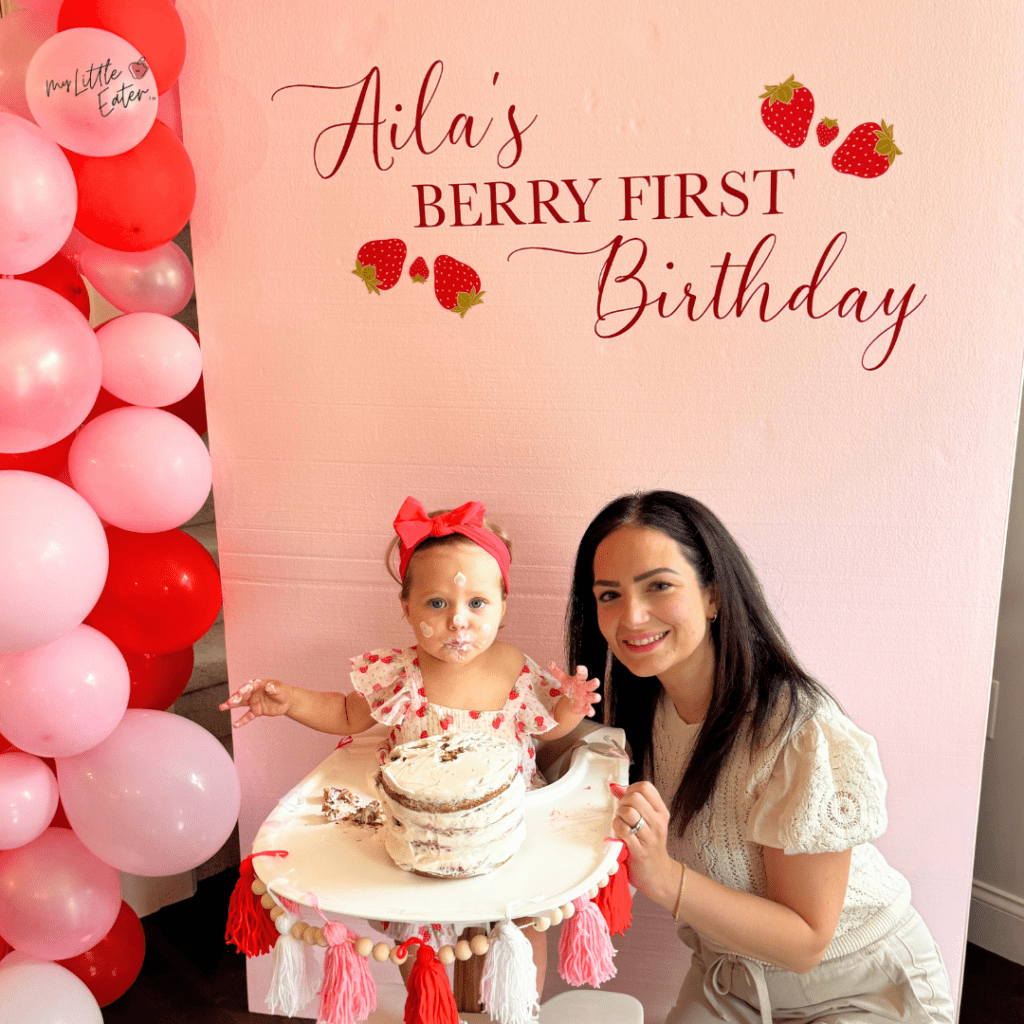 Aila's berry first birthday with baby Aila sitting in a high chair with her aunt, eating her multi-layer smash cake.