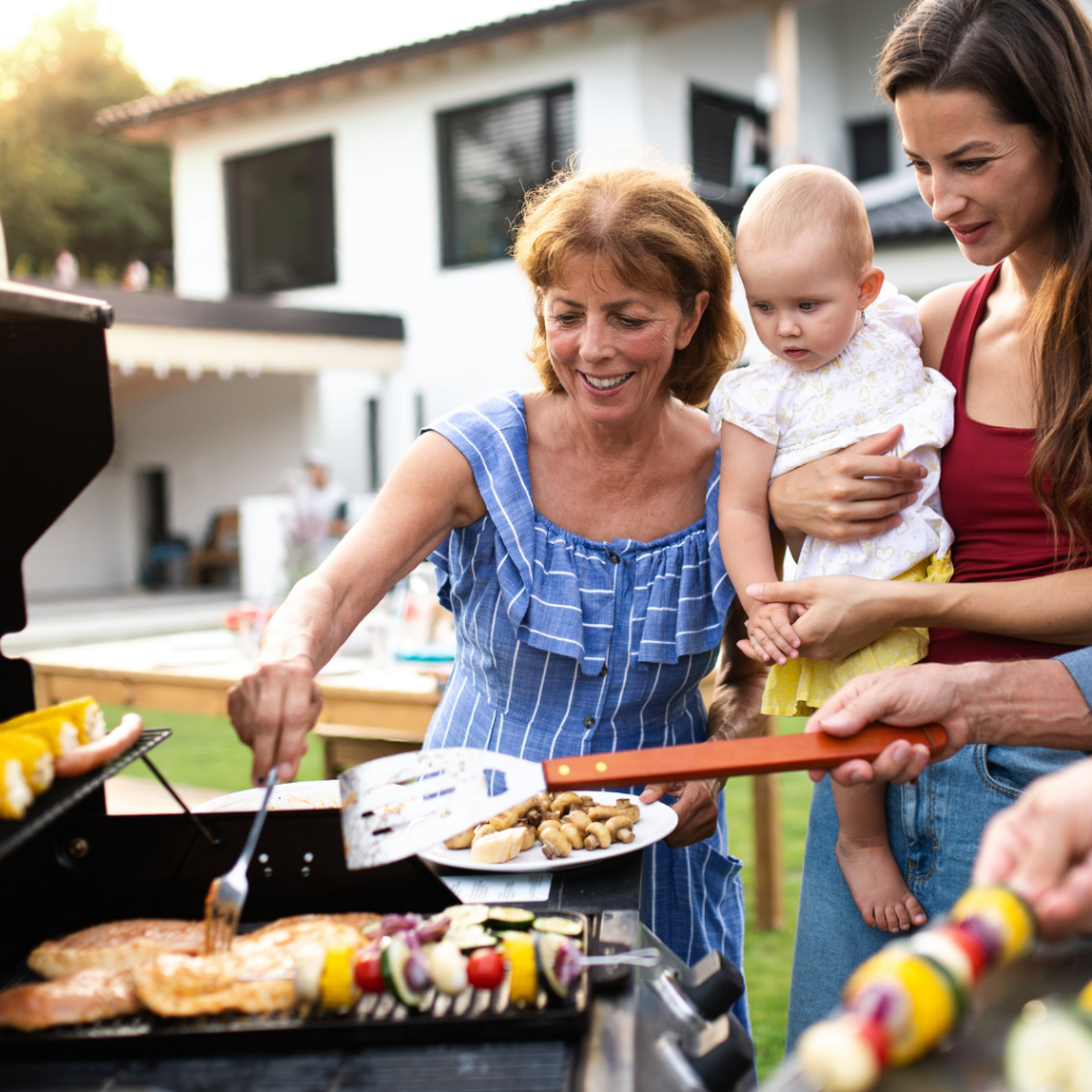 A family, including a baby under 12 months old, gathered around a barbecue while cooking skewers and chicken.