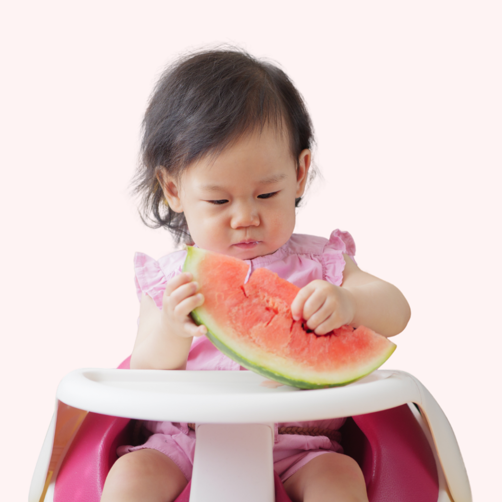 Baby girl sitting in a high chair holding up a large slice of sweet watermelon.