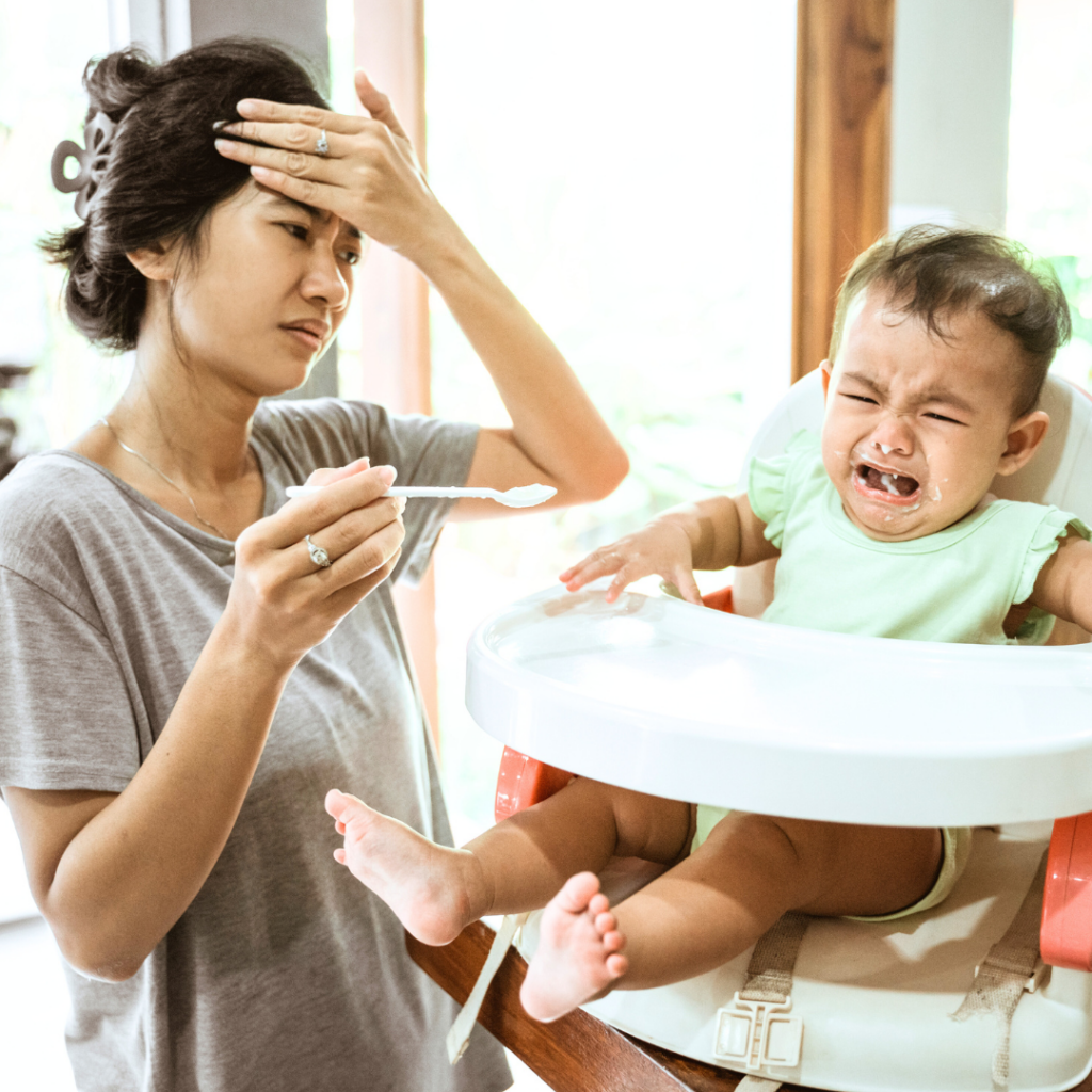 Mom trying to spoon feed baby who is sitting in a high chair; both appear frustrated.