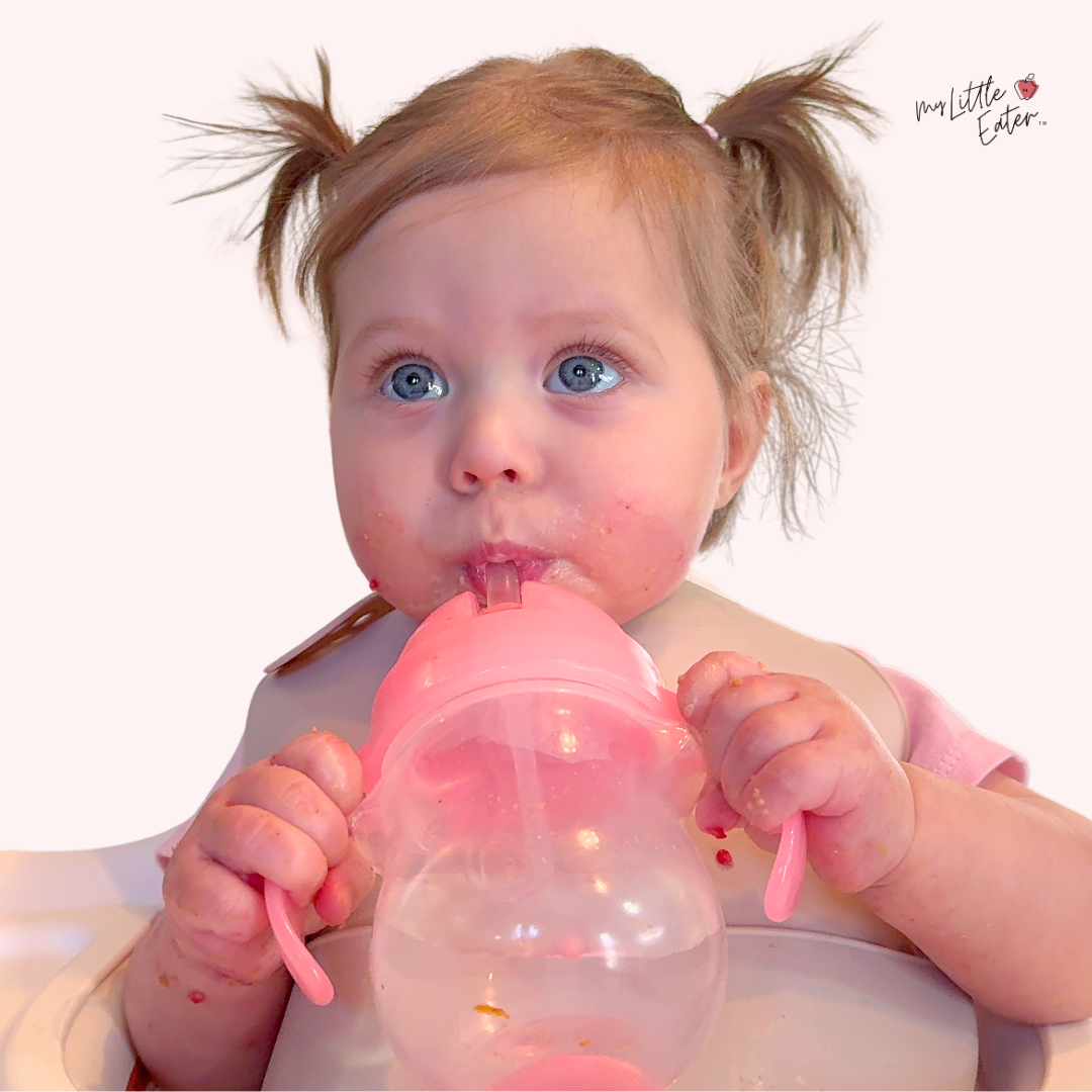 Aila sitting in her high chair drinking some water on her own from a pink munchkin straw cup. 