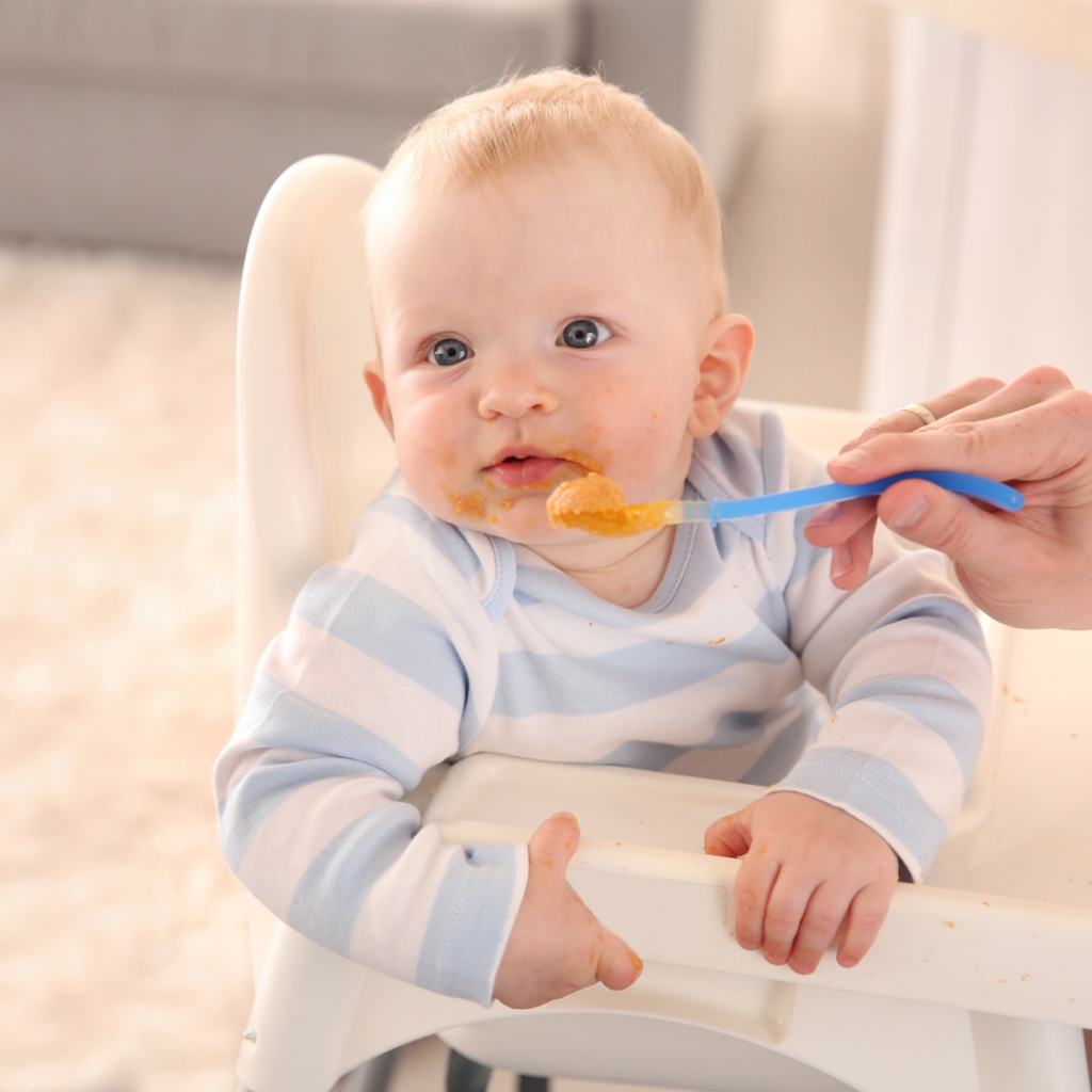 Parent trying to feed their baby just a little bit of pureed foods with a spoon while in their high chair, and baby has their head turned away.