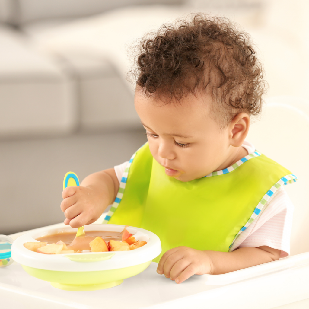 Young baby with good head control tries to feed themself solid food with a spoon by practicing scooping applesauce.