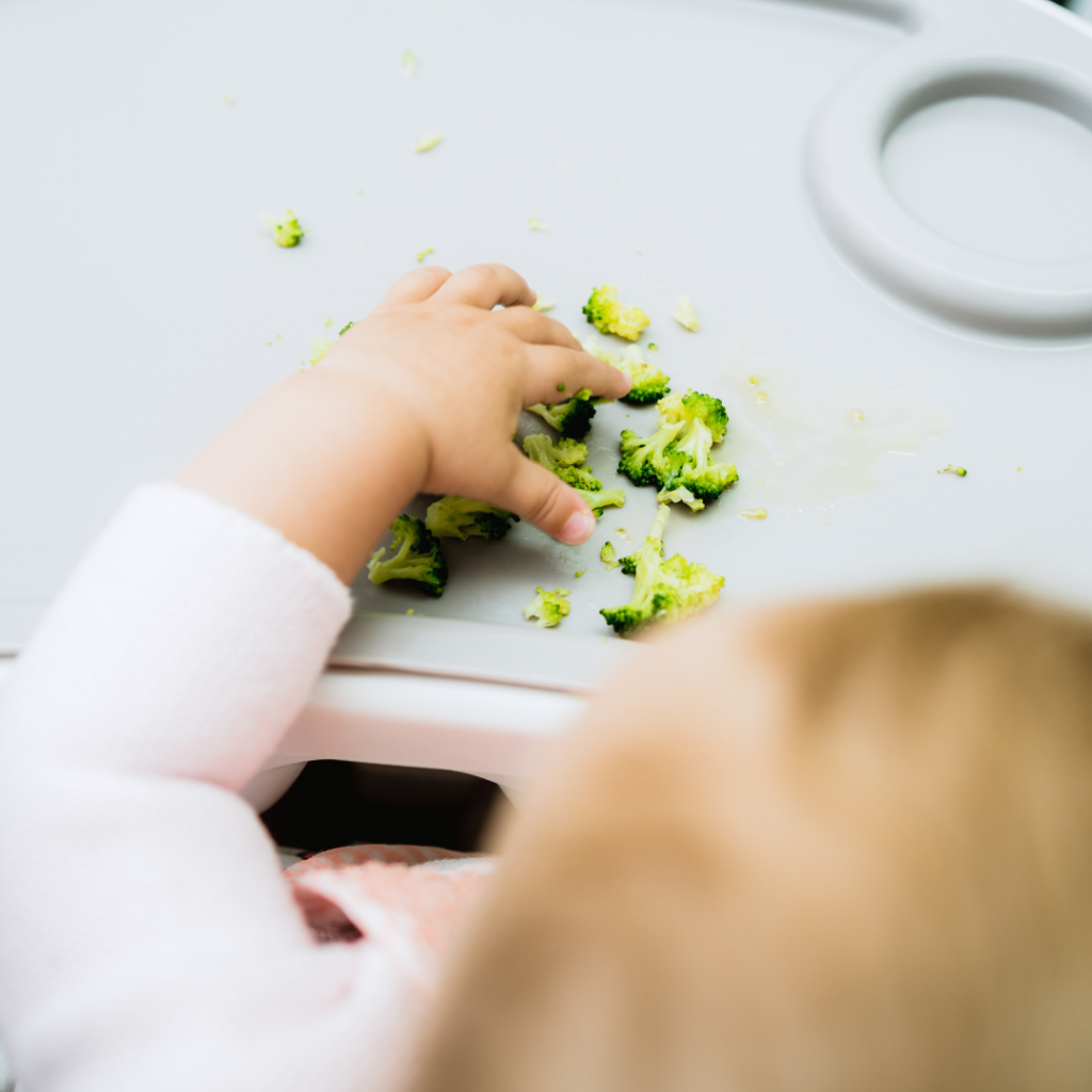 Overhead view of a baby sitting in a high chair reaching for florets of broccoli.