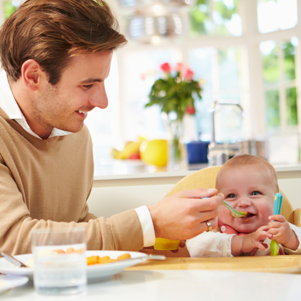 Father feeds his baby with a spoon at the dinner table.