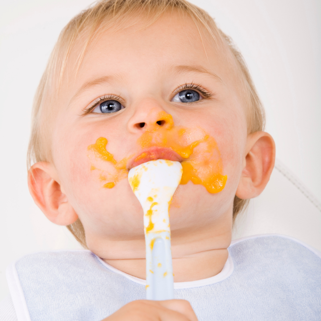 Baby self-feeding puree from a spoon with puree all over their face.